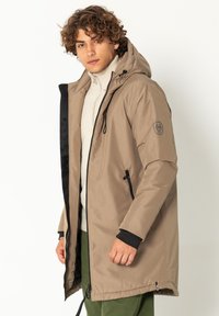 Hellbraune gepolsterte Jacke mit Kapuze, ausgestattet mit einem schwarzen Reißverschluss, Seitentaschen und einem Logo am Ärmel. Getragen über einem beigen Rollkragenpullover.