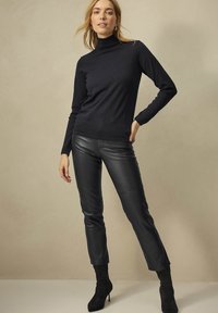 Schwarzer Rollkragenpullover aus weichem Stoff, kombiniert mit schmalen schwarzen Lederhosen. Das Modell trägt kniehohe schwarze Wildlederstiefel. Schlicher Hintergrund.