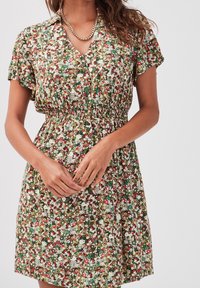 Femme portant une robe fleurie à manches courtes et taille élastique, dotée de boutons, accessoirisée d'un collier en chaîne doré, les mains jointes.