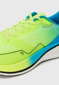 Sapatilha de corrida verde brilhante com parte superior em malha, cordões amarelos, detalhes azuis, padrão texturizado e sola branca com um degradê azul.