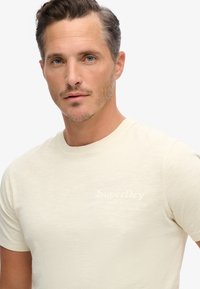 Camiseta de manga corta en beige claro, fabricada con una tela suave. Presenta un logotipo empotrado sutil en el pecho y un diseño clásico de cuello redondo.