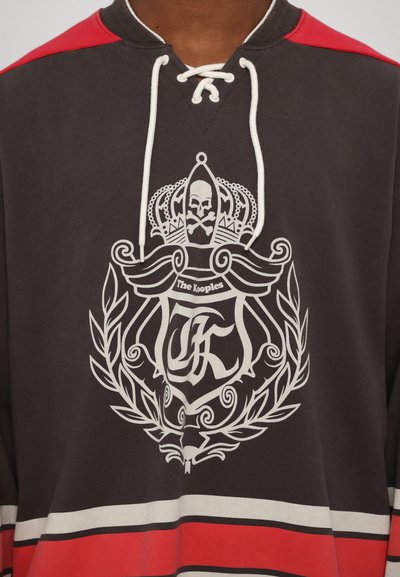 Camisa de manga larga gris oscuro con un gran diseño gráfico blanco que presenta una calavera, una corona y texto, acentuada con paneles y rayas rojas en los hombros.