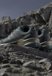 Ciemnozielone buty sportowe z siateczkową cholewką oraz szarymi akcentami, solidną podeszwą z bieżnikiem i teksturowaną powierzchnią. Widoczne logo na boku.