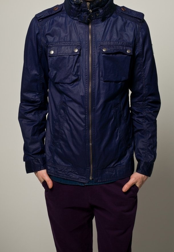 Veste bleu marine avec fermeture éclair à l'avant, col haut, deux poches poitrine et épaulettes, fabriquée à partir d'un tissu texturé.