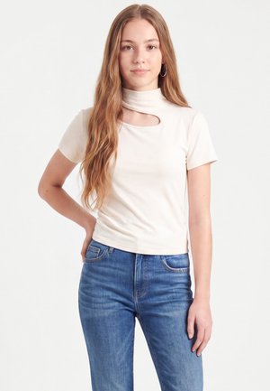 Jeune femme aux longs cheveux châtain clair portant un haut blanc à manches courtes avec une découpe près du cou et un jean bleu, debout avec une main sur la hanche.
