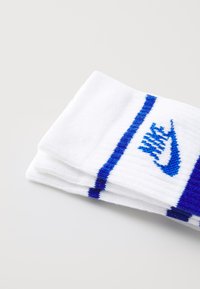 Chaussettes Nike, blanches avec des rayures bleu foncé, arborant un logo brodé bleu. Fabriquées en tissu texturé, pliées proprement pour l'affichage.