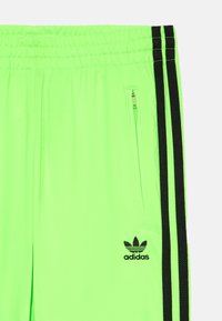 Neon zaļas sporta bikses ar melnām sānu strijām, elastīga josta un kabata ar rāvējslēdzi. Iekļauts neliels melns izšūts logs.