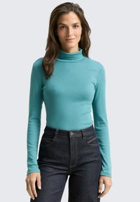 Langärmliges Turtleneck-Shirt aus türkisfarbenem Stoff mit einer figurbetonten Passform. Kombiniert mit einer dunklen Jeans im High-Waist-Stil und einem Knopfverschluss.