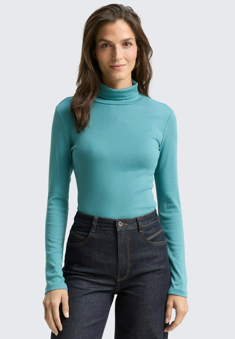 Langärmliges Turtleneck-Shirt aus türkisfarbenem Stoff mit einer figurbetonten Passform. Kombiniert mit einer dunklen Jeans im High-Waist-Stil und einem Knopfverschluss.