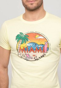 T-shirt en coton jaune avec un design graphique représentant un palmier, des vagues et le texte "MIAMI Sunset" en couleurs rouge et orange.