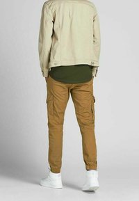 Bruna cargobyxor med sidfickor, elastiska mudd, matchade med en ljus beige jacka och en mörkgrön skjorta under, bärs med vita sneakers.