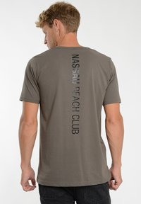 Gray cotton t-shirt with short sleeves, featuring vertical black text "NASSAU BEACH CLUB" down the center back. Soft texture, standard fit.