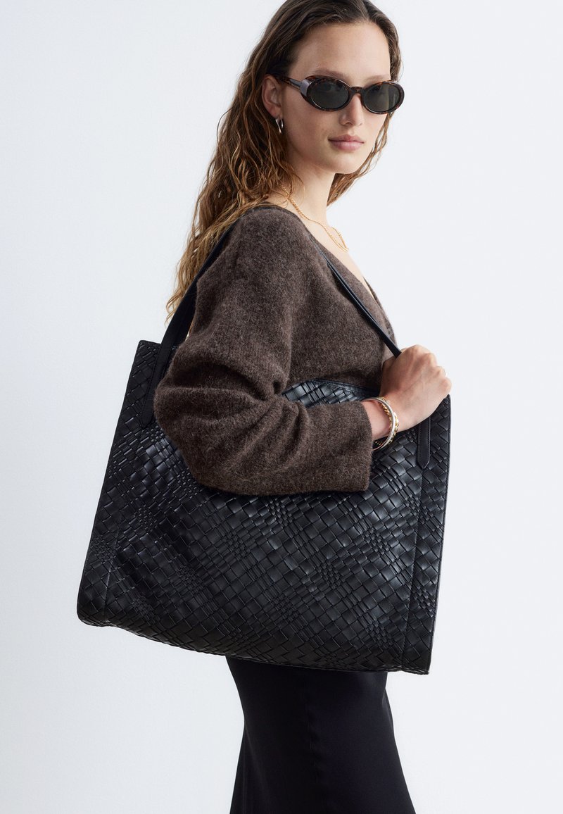 Femme portant des lunettes de soleil foncées et un pull marron, tenant un grand sac cabas en cuir noir tressé sur son épaule.