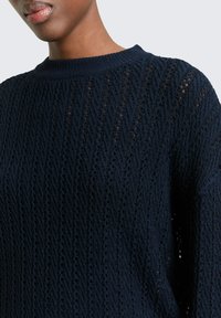 Close-up van een persoon die een marineblauwe gebreide trui draagt met een gestructureerd patroon en een geribde ronde halslijn.