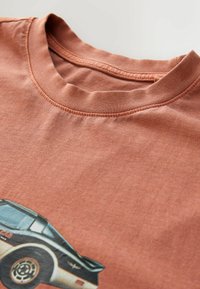 Rustfarvet bomulds tshirt med grafisk tryk af en sort bil. Blød tekstur, rund halsudskæring og synlige syningsdetaljer.