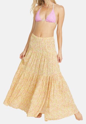 Frau trägt ein rosa gehäkeltes Bikinioberteil und einen langen, fließenden gelben Blumenrock mit Stufen und Knöpfen auf der Vorderseite.