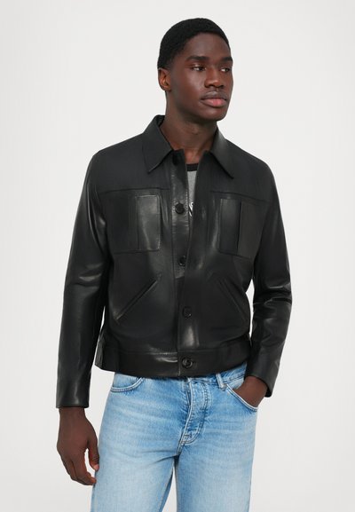 Sort læderjakke med knaplukning, to klaplommer og krave. Båret over en grå top og parret med lyseblå denimjeans.