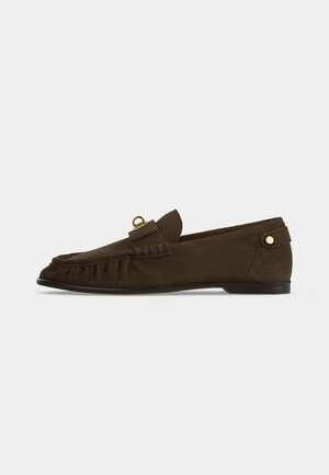 Mocasín de ante marrón oscuro con tacón bajo, costura lateral y detalle de aro metálico y botón dorado sobre un fondo liso.