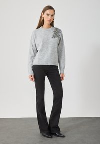 Suéter gris de punto con mangas largas, adornado con bordados florales en el hombro izquierdo, combinado con jeans de campana negros y botas negras hasta el tobillo.