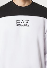 Personne portant un sweat-shirt noir et blanc avec le logo "EA7 Emporio Armani" imprimé sur la poitrine.
