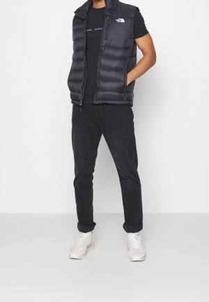 Mand stående med hænderne i lommerne iført sort dunvest, sort t-shirt, sorte jeans og hvide sneakers mod en ensfarvet baggrund.