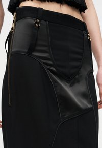 Detailný záber na čiernu sukňu s lesklým panelom pripomínajúcim kožu, zlatým zipsom na boku a nastaviteľnými ramienkami so zlatými kovaniami v páse.