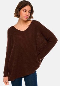 Pull en tricot marron avec une coupe décontractée et oversized, un large col rond et des manches longues ornées de motifs texturés. Associé à un jean bleu.