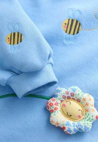 Modrá látka se vyšívanými usmívajícími se včelami a barevnou nášivkou usmívající se květiny s květinovými vzory u manžety rukávu.