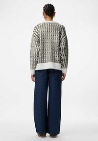 Pull en maille gris clair avec un motif en chaîne vert foncé, poignets blancs, et une coupe ample, associé à un jean large bleu foncé.