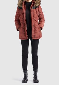 Fodrad röd jacka med pälsfoder på huvan och svarta läderdetaljer, med dragkedja fram, sidofickor och kviltat mönster. Svarta leggings och stövlar.