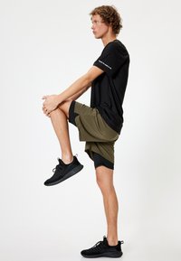 Camiseta negra de manga corta combinada con pantalones cortos verde oliva con acentos negros. Zapatillas deportivas negras completan el conjunto.