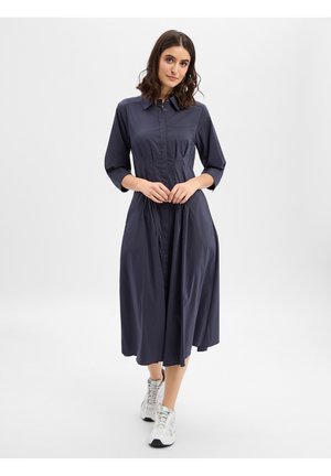 Kobieta stojąca w granatowej, długorękawowej, zapinanej sukience midi, w białych trampkach na jasnym, jednolitym tle.