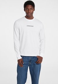 Langærmet hvid bomulds skjorte med "GUESS JEANS" tekst i sort. Kombineret med blå denim jeans for et afslappet look.
