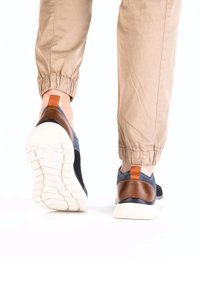 Les baskets présentent un dessus en mesh bleu marine avec des accents en cuir marron, une semelle en caoutchouc blanche et une languette à l'arrière pour un enfilage facile.