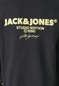 Camisa de algodón negra de manga larga con un gran texto amarillo que dice "JACK & JONES STUDIO EDITION ©1990" y un gráfico de firma en la parte inferior.