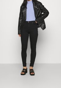 Mujer con chaqueta de cuero negra, suéter de canalé púrpura claro, pantalones skinny negros y sandalias de tacón negro con hebillas doradas.