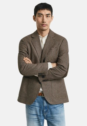 Mann steht mit verschränkten Armen, trägt ein braunes karriertes Sakko, ein weißes Hemd und eine blaue Jeans vor einem schlichten weißen Hintergrund.