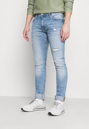 Person iført lyseblå, slidte skinny jeans med opbuk ved anklerne, kombineret med hvide sneakers og en grøn langærmet top.