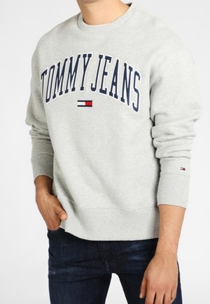 Mężczyzna noszący jasnoszarą bluzę Tommy Jeans z granatowym napisem i logo, w połączeniu z ciemnoniebieskimi jeansami, ręka w kieszeni.