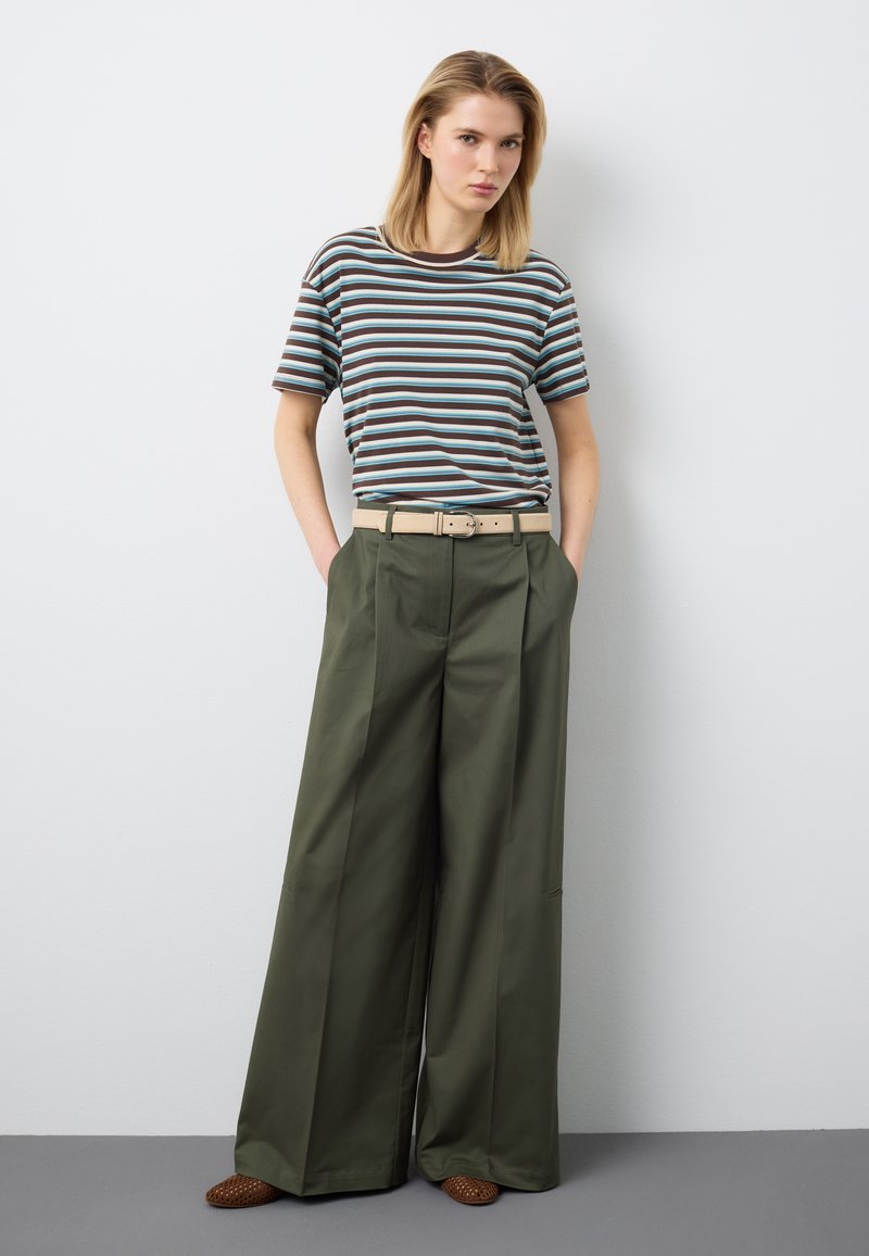 Junge Person in einem gestreiften braunen, blauen und weißen T-Shirt mit beigem Gürtel, weiten olivgrünen Hosen und braunen gewebten Schuhen, die vor einer weißen Wand steht.