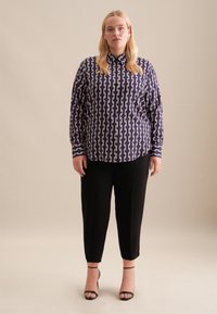 Blusa viola a maniche lunghe con motivo a catena bianca, colletto a camicia, abbinata a pantaloni neri sartoriali e tacchi a strappy neri.