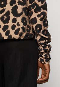 Blouse imprimée léopard beige et noir avec de longues manches froncées, associée à un pantalon en tissu noir. Texture lisse, design court.