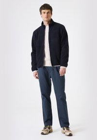Chaqueta de forro polar navy con cremallera en la parte delantera, cuello alto y bolsillo en el pecho, combinada con un suéter crema, pantalones azules y zapatillas beige.