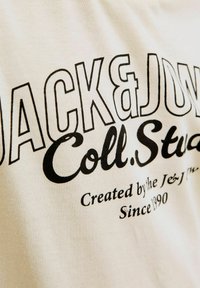 Krēmīgi balts kokvilnas T-krekls ar drosmīgu melnu tipogrāfiju. Teksts ietver "JACK & JONES Coll. Studio" un "Izveidots no J&J... Kopš 1990. gada."