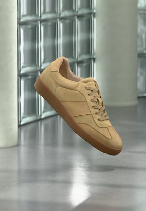 Beżowy zamszowy sneaker z gumową podeszwą, wiązany, unoszący się wewnątrz na polerowanej posadzce betonowej obok ściany z bloków szklanych.