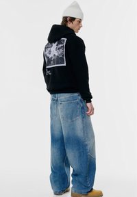 Felpa nera con stampa grafica sul retro, abbinata a jeans di denim chiaro e larghi e stivali da lavoro color sabbia. Berretto di cotone bianco indossato.
