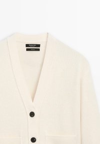 Cardigan in lana a coste color crema con scollo a V, bottoni neri e tasca a sinistra. Le etichette indicano il materiale e il nome del marchio.