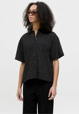 Chemise noire à manches courtes avec un design en œillets floraux, boutonnée devant et coupe décontractée. Associée à un pantalon noir assorti.