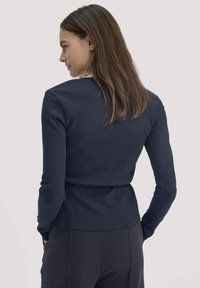 Vrouw met lang bruin haar draagt een strak navy langarmshirt en donkere broek, staat met haar handen in de zakken en kijkt naar links.