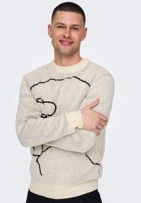 Pletený krémový svetr s černým abstraktním vzorem, vroubkovaným výstřihem a manžetami, s volným střihem a měkkou texturou.